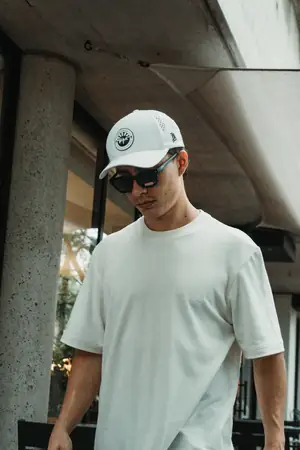 Con gorra y lentes de sol, polera blanca UEBOS bajo un techo de concreto